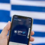 Χέρι που κρατά smartphone με ανοιχτή την εφαρμογή Gov.gr Wallet και φόντο την ελληνική σημαία θολωμένη