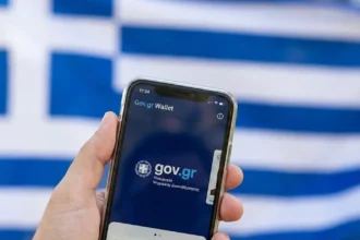 Χέρι που κρατά smartphone με ανοιχτή την εφαρμογή Gov.gr Wallet και φόντο την ελληνική σημαία θολωμένη