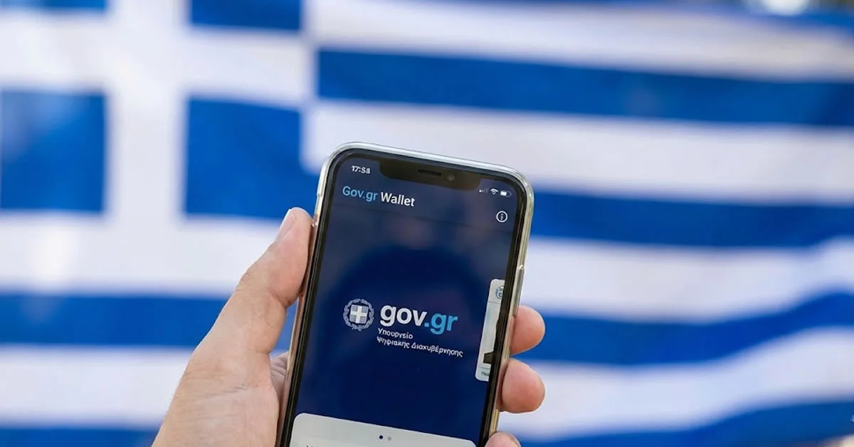 Χέρι που κρατά smartphone με ανοιχτή την εφαρμογή Gov.gr Wallet και φόντο την ελληνική σημαία θολωμένη