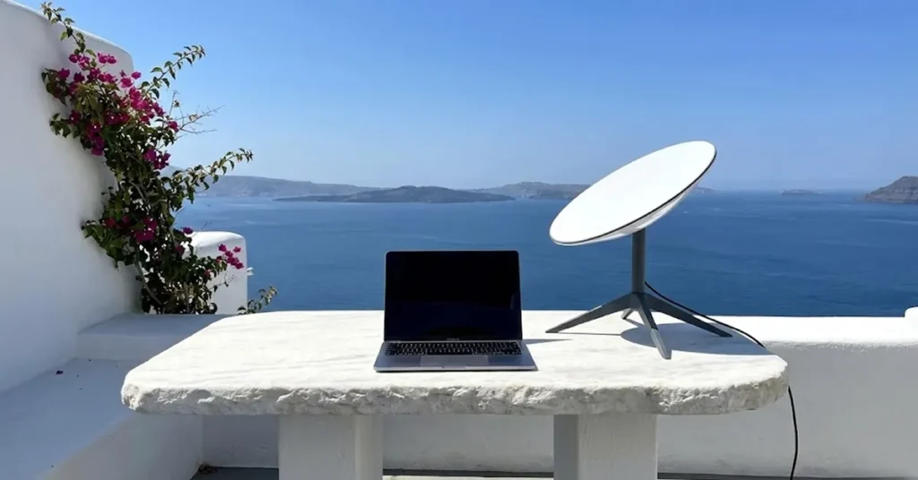 δείχνει ένα λευκό κυκλαδίτικο τραπέζι με ένα laptop, έχοντας στο πλάι το δορυφορικό πιάτο του Starlink και στο βάθος το απέραντο γαλάζιο, συμβολίζοντας την απόλυτη ελευθερία εργασίας